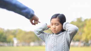 Ein Kind steht draußen auf einer Wiese, die Hände an den Ohren, mit einem genervten oder traurigen Gesichtsausdruck. Eine ausgestreckte Hand einer erwachsenen Person deutet auf das Kind, als würde es zurechtgewiesen werden.
