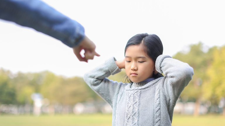 Ein Kind steht draußen auf einer Wiese, die Hände an den Ohren, mit einem genervten oder traurigen Gesichtsausdruck. Eine ausgestreckte Hand einer erwachsenen Person deutet auf das Kind, als würde es zurechtgewiesen werden.