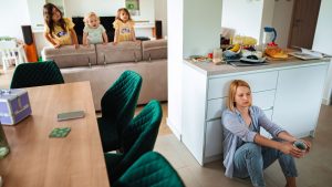 Mutter sitzt mit einer Tasse auf dem Boden in der Küche, während drei kleine Kinder sie beobachten; Küche und Essbereich wirken unaufgeräumt.