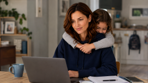 Eine lächelnde Mutter arbeitet am Laptop in einem Büro, während ihr kleines Kind sich liebevoll von hinten an sie klammert – Symbol für die Balance zwischen Beruf und Familie.