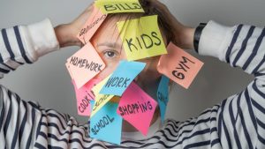Frau mit bunten Klebezetteln im Gesicht, beschriftet mit Aufgaben wie „Kids“, „Work“, „Shopping“ – Symbol für Mental Load im Familienalltag.