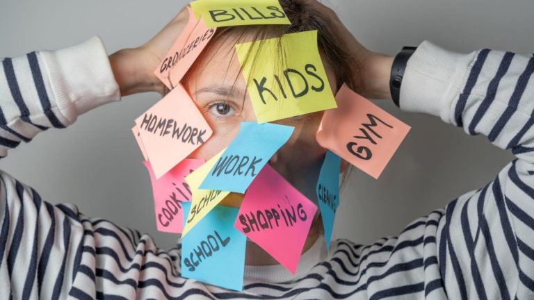 Frau mit bunten Klebezetteln im Gesicht, beschriftet mit Aufgaben wie „Kids“, „Work“, „Shopping“ – Symbol für Mental Load im Familienalltag.